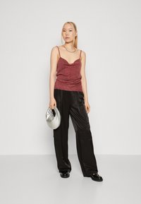 Bordeaux metallic top met dunne bandjes, gecombineerd met zwarte satijnen broek met wijde pijpen. Model houdt een zilveren clutch vast en staat tegen een effen achtergrond.