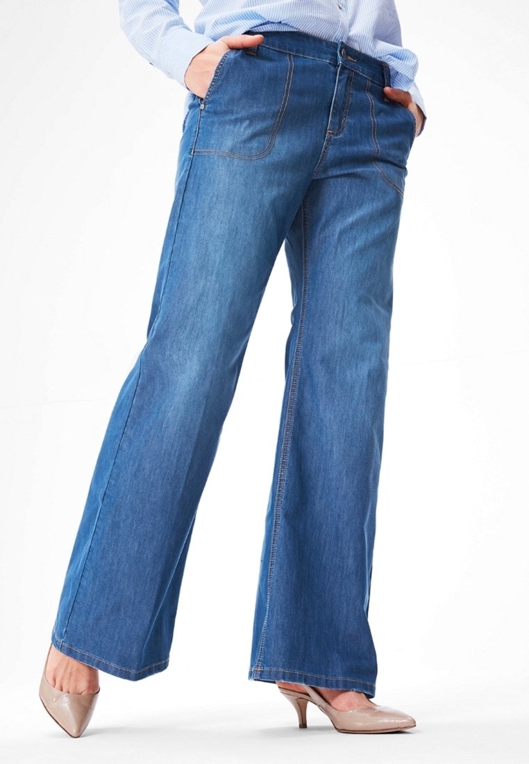 Personne portant un jean bleu à jambes larges, une chemise rayée bleu clair, les mains dans les poches et des talons beiges sur un fond blanc uni.