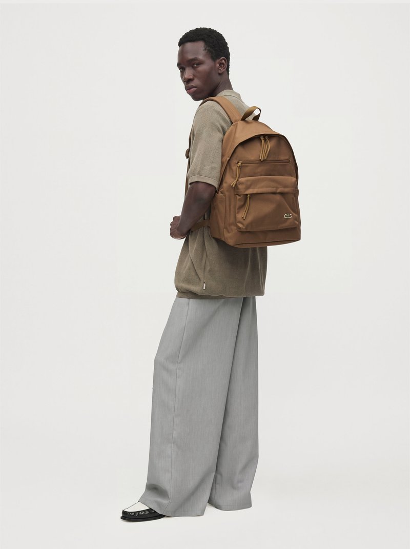 Un homme regarde par-dessus son épaule tout en présentant un sac à dos marron, un haut beige, un pantalon gris ample et des chaussures noires sur un fond clair uni.