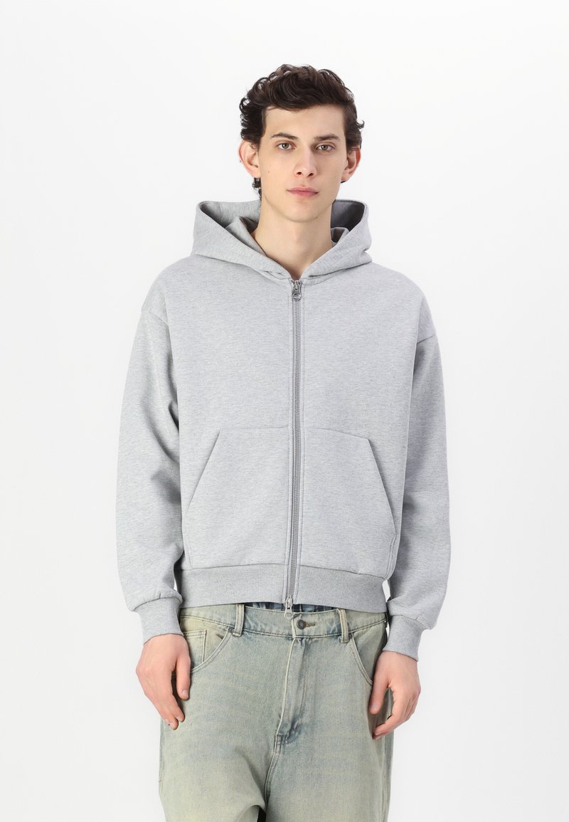 Sweat à capuche gris zippé en tissu doux, avec une grande capuche, deux poches avant et des poignets et un ourlet côtelés. Porté sur un jean large.