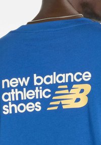 T-shirt in cotone blu con testo bianco e dorato: "scarpe da atletica New Balance." Design semplice con collo tondo e una texture liscia.