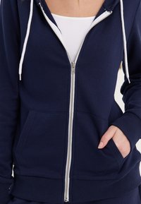 Mornarska zip-up dukserica od mekog materijala, s bijelim vezicama, prednjim džepovima i rebrastim manžetama i donjim rubom. Srebrni detalj na patentnom zatvaraču.