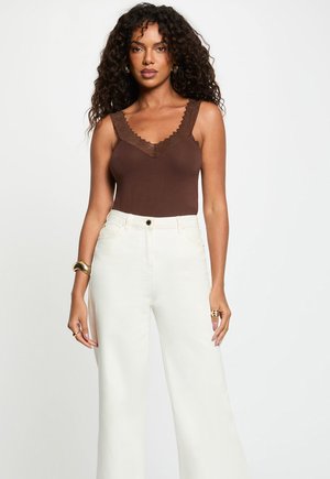 Vrouw met krullend haar, die een bruin mouwloos topje draagt met kanten afwerking en witte, wijde, hooggetailleerde broek, staand tegen een effen achtergrond.