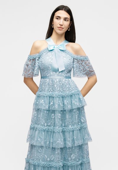 Γυναίκα φορούν ανοιχτό γαλάζιο δαντελένιο φόρεμα με σχεδιασμό που αφήνει ακάλυπτους τους ώμους, κοντά διαφανή μανίκια, στρωματοποιημένες βολάν και μεταξωτό φιόγκο στο ντεκολτέ.