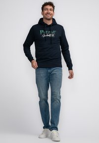Granatowy hoodie z grafika na przodzie "PetroD Industries", z kapturem na sznurku, noszony z jasnoniebieskimi dżinsami i białymi trampkami. Model stoi i się uśmiecha.