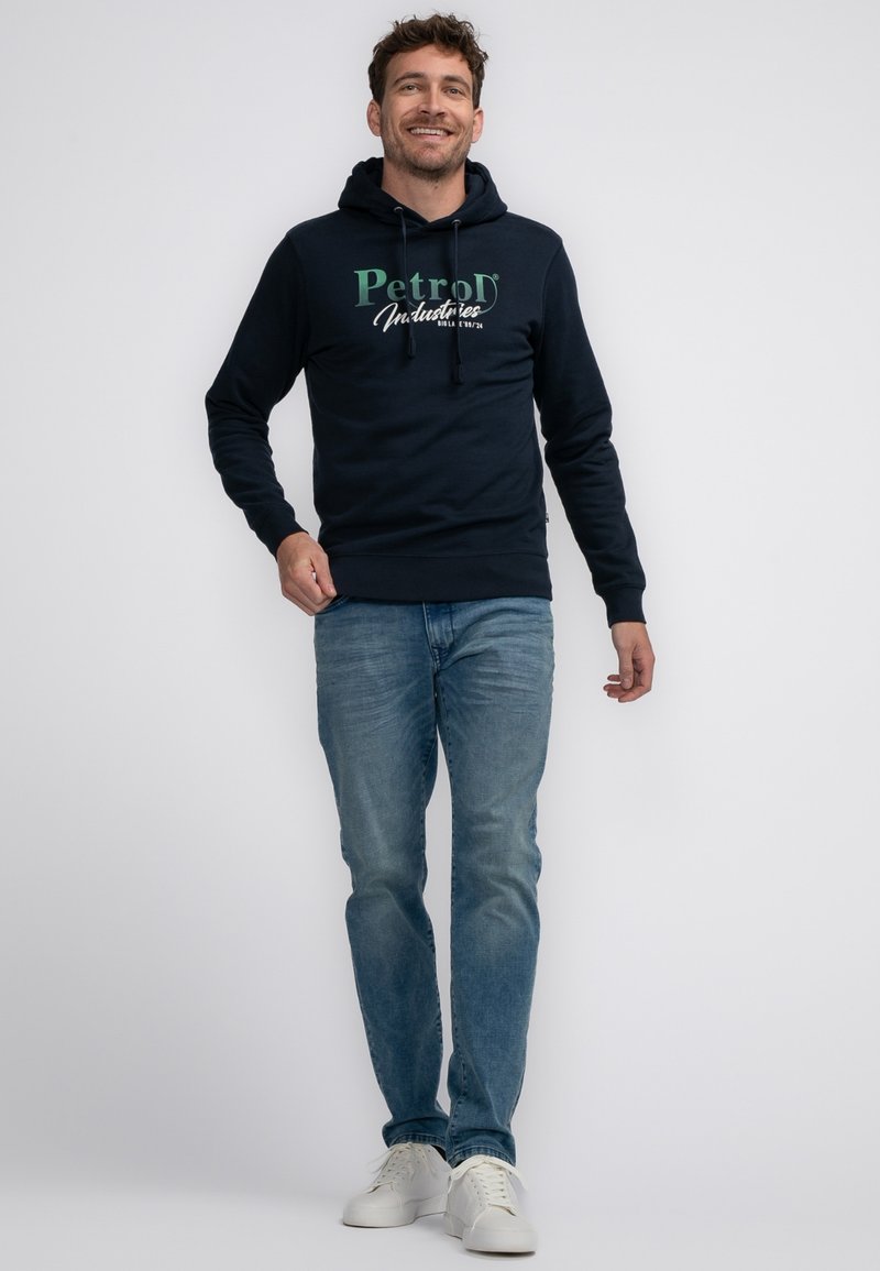 Granatowy hoodie z grafika na przodzie "PetroD Industries", z kapturem na sznurku, noszony z jasnoniebieskimi dżinsami i białymi trampkami. Model stoi i się uśmiecha.
