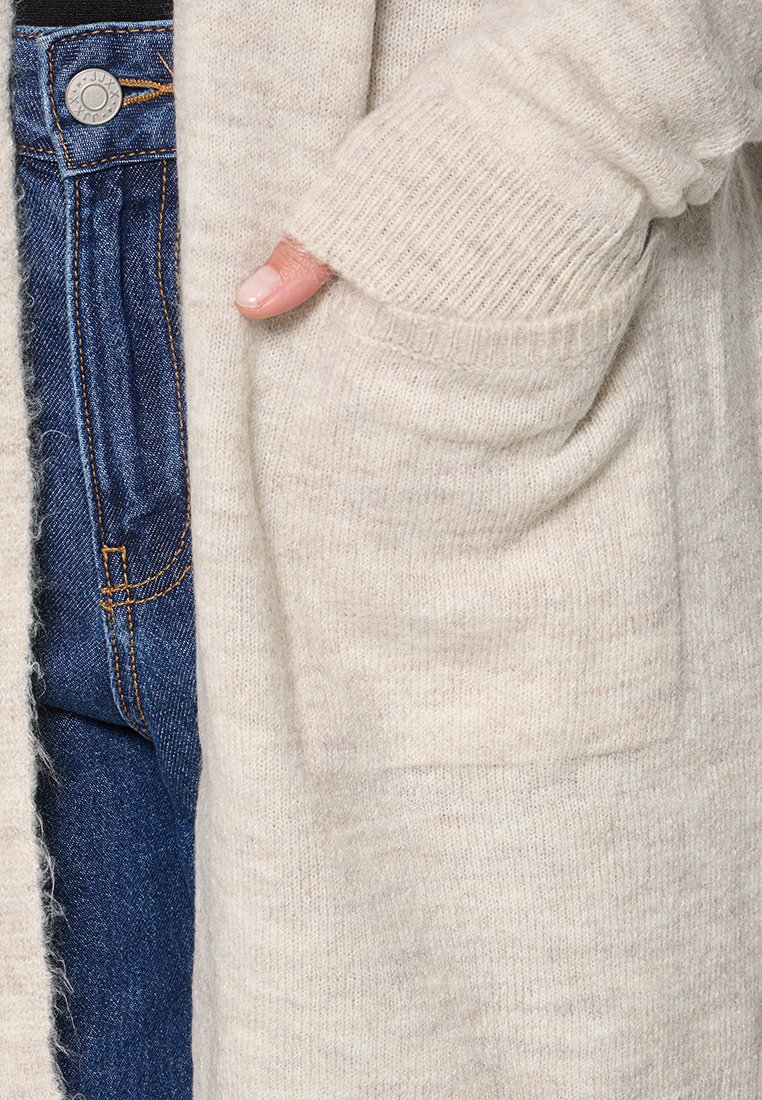 Lichtbeige cardigan met geribde manchetten, zachte textuur en een zak. Draag het over donkerblauwe spijkerbroeken met zichtbare stiksels en een knoop.