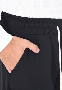 Mano che riposa all'interno della tasca dei pantaloni neri in tessuto elasticizzato con cordoncino bianco, abbinati a una maglietta bianca a maniche lunghe a coste.