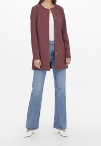 Mörkröd lång kappa med rak skärning, tryckknappar och rund halsringning. Bärs över en vit topp och matchas med ljusblå jeans med utställda ben.