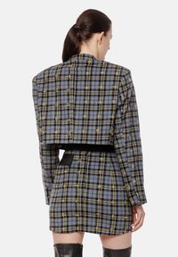 Kobieta z ciemnym kucykiem, ubrana w dopasowaną szaro-skalną kurtkę z kratą i mini spódnicę z żółtymi akcentami, połączona z czarnymi kozakami do kolan.