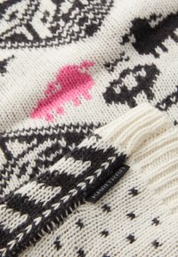 Strikkegenser med svart og hvitt mønster, med en rosa aksent. Laget av ull, med ribbestrikket mansjett og merke på kanten.