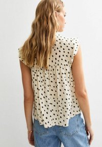 Cream-colored sleeveless top with black polka dots, ruffled shoulders, and a loose fit, paired with light blue denim jeans.