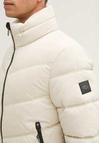 Chaqueta tipo plumón en color crema claro, con un cuello alto, costuras horizontales, bolsillos laterales con cremallera y un parche de logo negro en la manga.