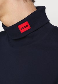 Námořnický rolák s texturovanou tkaninou, s červeným obdélníkovým štítkem, na kterém je černě vytištěno "HUGO". Detailní pohled zdůrazňuje design límce.