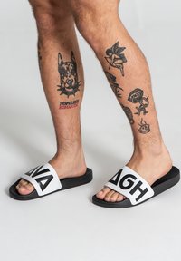 Chanclas de slide en blanco y negro con letras negras en negrita en una tira ancha, fabricadas con un material suave y duradero. Diseño de suela plana.