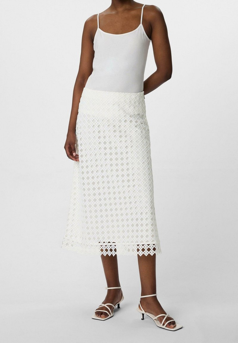 Jupe midi en dentelle blanche avec un motif de losanges et un ourlet festonné, associée à un débardeur blanc ajusté et des sandales à lanières.