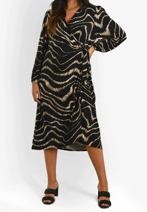 Vrouw draagt een zwarte wikkeljurk met beige abstracte golfpatronen, lange mouwen en zwarte open-toe pumps, staand met één hand op de heup.