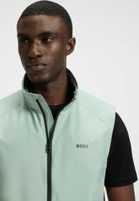 Hellgrüne ärmellose Weste mit hohem Kragen und schwarzem Reißverschluss. Hat ein dezentes strukturiertes Muster und ein graues "BOSS"-Logo auf der Brust.
