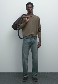 Ung mand iført brun zip-up polo, falmede blå jeans, mørke sko, med en stor sort taske over skulderen mod en ensfarvet grå væg.