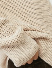 Maglione beige in maglia con un motivo testurizzato, polsini a coste e un tessuto morbido e flessibile, mostrato con parte di una mano che afferra il tessuto.