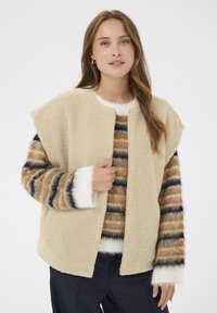 Bežičasti, teksturirani flis kaput s otvorenim prednjim dijelom i širokim ramenima. Uparen s prugastim, višebojnom pletenom puloverom s bijelim detaljima.