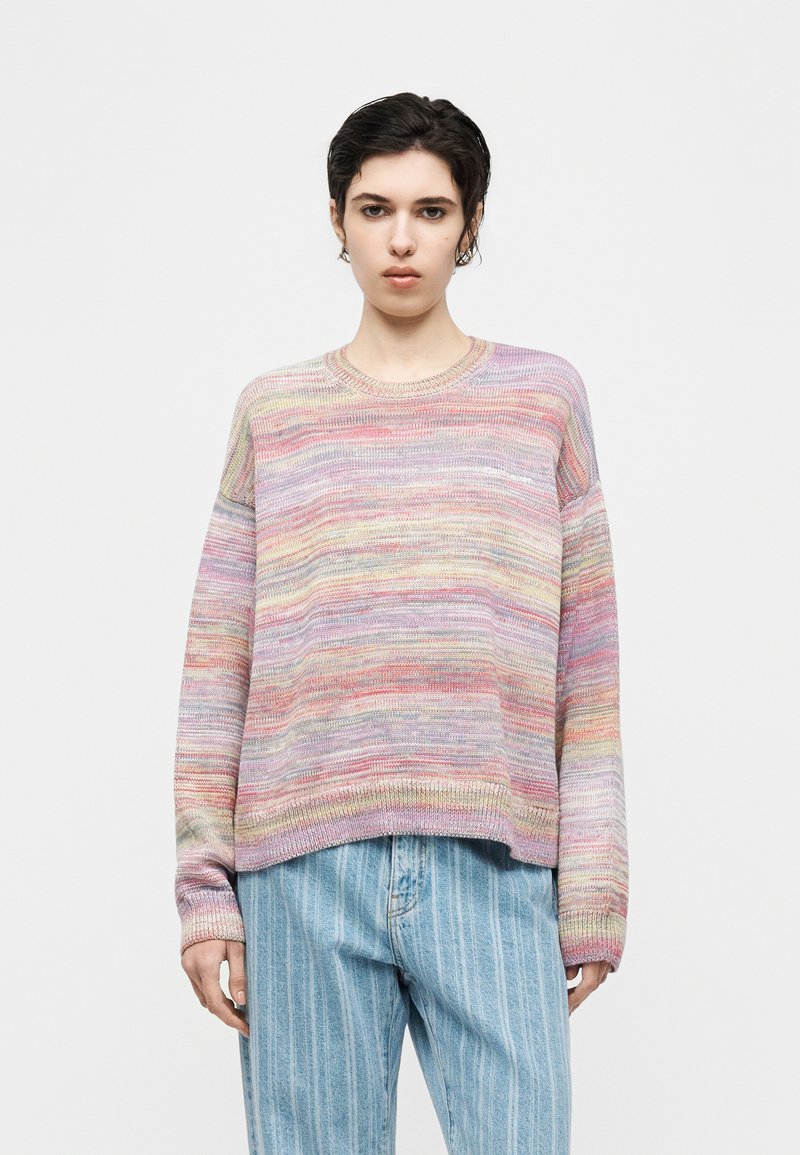 Persona giovane con capelli corti e scuri che indossa un maglione a righe multicolore e jeans larghi azzurri chiari su uno sfondo bianco semplice.