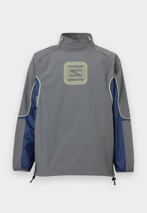 Veste grise avec des panneaux latéraux bleu marine et une bordure jaune néon, présentant un logo géométrique centré sur le haut du dos et des fermetures éclair sur le col.