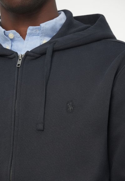 Ναυτικό φούτερ με φερμουάρ σε μαλακό ύφασμα με κορδόνια, διαθέτοντας ένα μικρό γυαλισμένο λογότυπο στο στήθος και μια ανοιχτό μπλε πουκάμισο από κάτω.