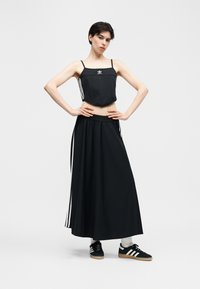 Junge Person steht mit Händen in die Hüften gestemmt, trägt ein schwarzes Adidas-Trägershirt, langen schwarzen Rock mit weißen Streifen, weiße Socken und schwarze Turnschuhe.