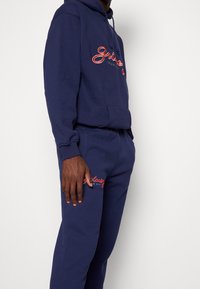 Marinblå hoodtröja och sweatpants-set, med broderad logotyp i röd. Tillverkad av mjukt, texturerat tyg med en framficka och dragsko på huvan.