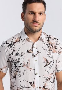 Chemise à manches courtes, blanche, avec un motif floral et d'oiseaux en rose et noir. Elle possède un col à boutons et des boutons visibles.