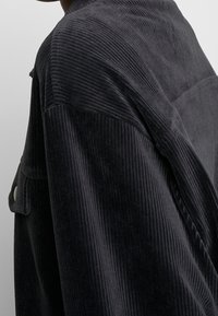 Camisa-chaqueta de pana negra con canalé vertical, que presenta un collar estructurado, bolsillo frontal y detalle de botones en la manga.