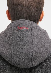 Grijze wollen capuchon met zichtbare textuur en stiksels. Bevat een klein rood geborduurd label aan de achterkant. Vouwen en naden zijn prominent aanwezig.