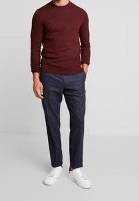 Bordeauxrode gebreide trui met lange mouwen, gecombineerd met donkergrijze getextureerde broek en witte sneakers, nonchalant gedragen met één hand in de zak.