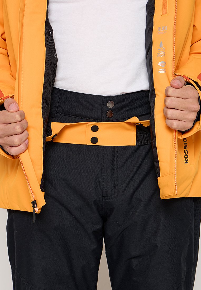 Osoba w czarnych spodniach i żółtej kurtce wodoodpornej oraz wiatroszczelnej, zapinająca zamek kurtki z widocznym logo oraz funkcjonalnymi detalami.