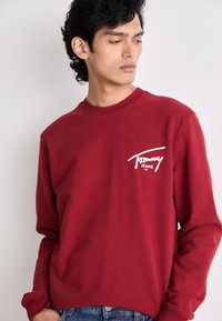 Giovane uomo che guarda a sinistra, indossando una felpa rossa di Tommy Jeans e jeans blu su uno sfondo chiaro e semplice.