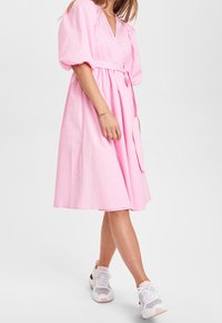 Rosa bomullsklänning med djup V-ringning och puffärmar, bältad midja, A-linjeformad kjol, kombinerad med vita och grå sneakers.