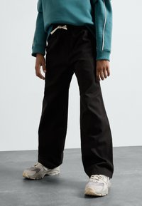 Pantalons larges noirs avec une taille à cordon et une finition en coton texturé, associés à des baskets grises et un sweat-shirt teal.