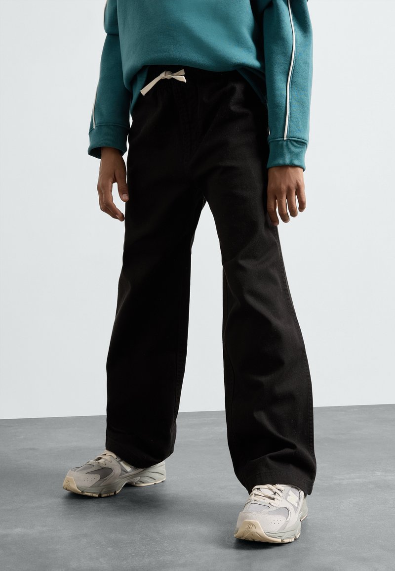 Pantalons larges noirs avec une taille à cordon et une finition en coton texturé, associés à des baskets grises et un sweat-shirt teal.