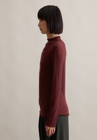 Burgunderfarbener, gerippter Langarmoberteil mit einem Rüschensaum am Ausschnitt und an den Säumen, schmal geschnitten und aus strukturiertem Stoff. Seitenansicht gezeigt.