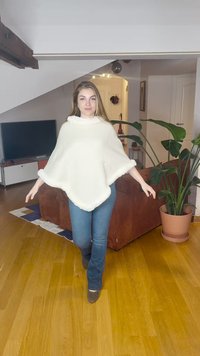 Valge poncho pehme tekstuuriga, millel on laineline ääris ja karvane ääris, kantud koos siniste teksade ja pruuni jalanõudega hubases siseruumis.