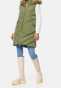 Gevoerde olijfgroene vest met een bontomrand capuchon, voorzien van een ritsluiting en zijzakken; gecombineerd met lichtblauwe jeans en beige kniehoge laarzen.
