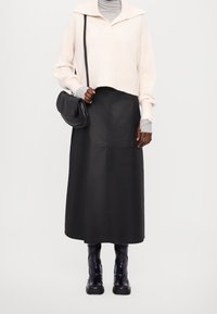 Schwarzer Leder-Midirock mit vorderen Nahtdetails, kombiniert mit einem hellbeigen, gestrickten Cropped-Pullover. Accessoires umfassen eine schwarze Schultertasche.