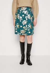 Teal skjørt med gullblomstermønster, knelangt, kombinert med knelangne svarte skinnstøvler. Strikket beige genser er hatt på toppen.