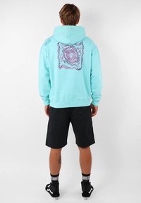 Haut à capuche turquoise avec un graphisme texturé violet dans le dos. Mélange de coton, ourlets côtelés aux poignets et en bas. Associé avec des shorts noirs et des baskets.