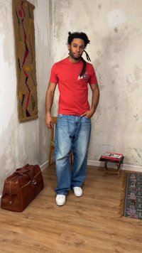 Άνδρας με dreadlocks στέκεται σε εσωτερικό χώρο φορώντας κόκκινο μπλουζάκι, φαρδιά τζιν και λευκά αθλητικά παπούτσια, δίπλα σε καφέ δερμάτινη τσάντα και ξύλινη καρέκλα.