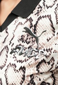 Πόλο μπλούζα με μαύρο γιακά, με σχέδιο δέρματος φιδιού σε κρεμ και καφέ αποχρώσεις, και ασημένιο λογότυπο Puma με τη λέξη "AWIDE" από κάτω.