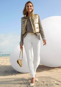 Goldene Daunenweste über einem hellgrauen Strickpullover, weißen Slim-Fit-Hosen und einer kleinen beigen Tasche. Sand und geometrische Formen im Hintergrund.