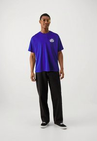 Violets kokvilnas T-krekls ar īsām piedurknēm un grafisku detaļu kreisajā krūtīs. Kombinēts ar melnām, brīva piegriezuma džinsa biksēm un melniem sporta apaviem.