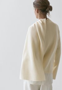 Cream-colored wool sweater with a draped design, featuring an open back and flowing sleeves, worn over a white layer.